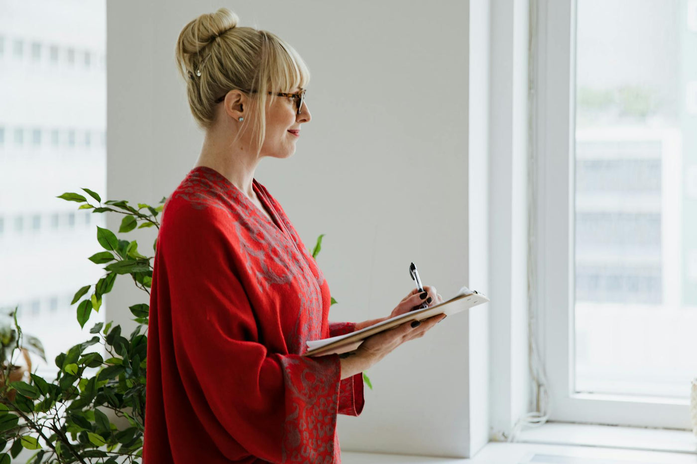 Profile of a woman in a red shawl indoors, holding a clipboard, near a window.