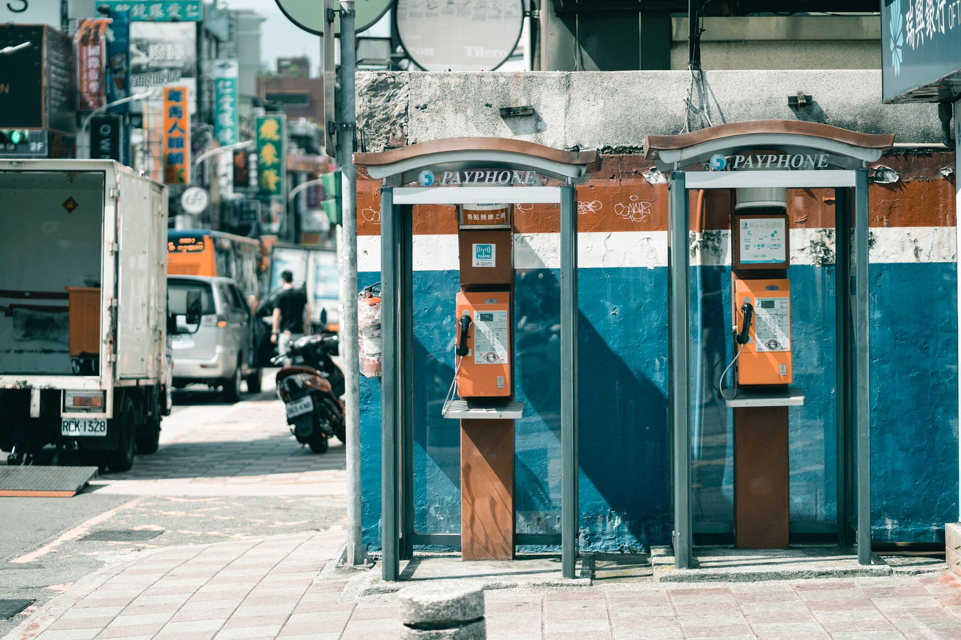 Identical aged call boxes with hanging telephones near rough concrete wall with shabby surface on pavement near asphalt roadway with driving vehicles and parked motorcycle in town in daylight