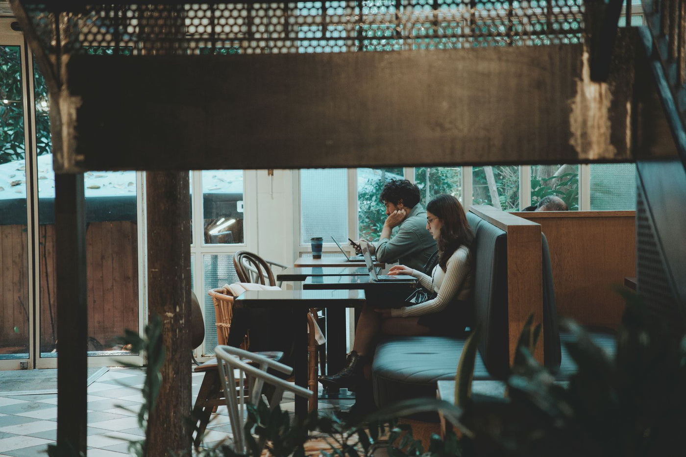 Two young adults working on laptops in a stylish, cozy café interior surrounded by plants.
