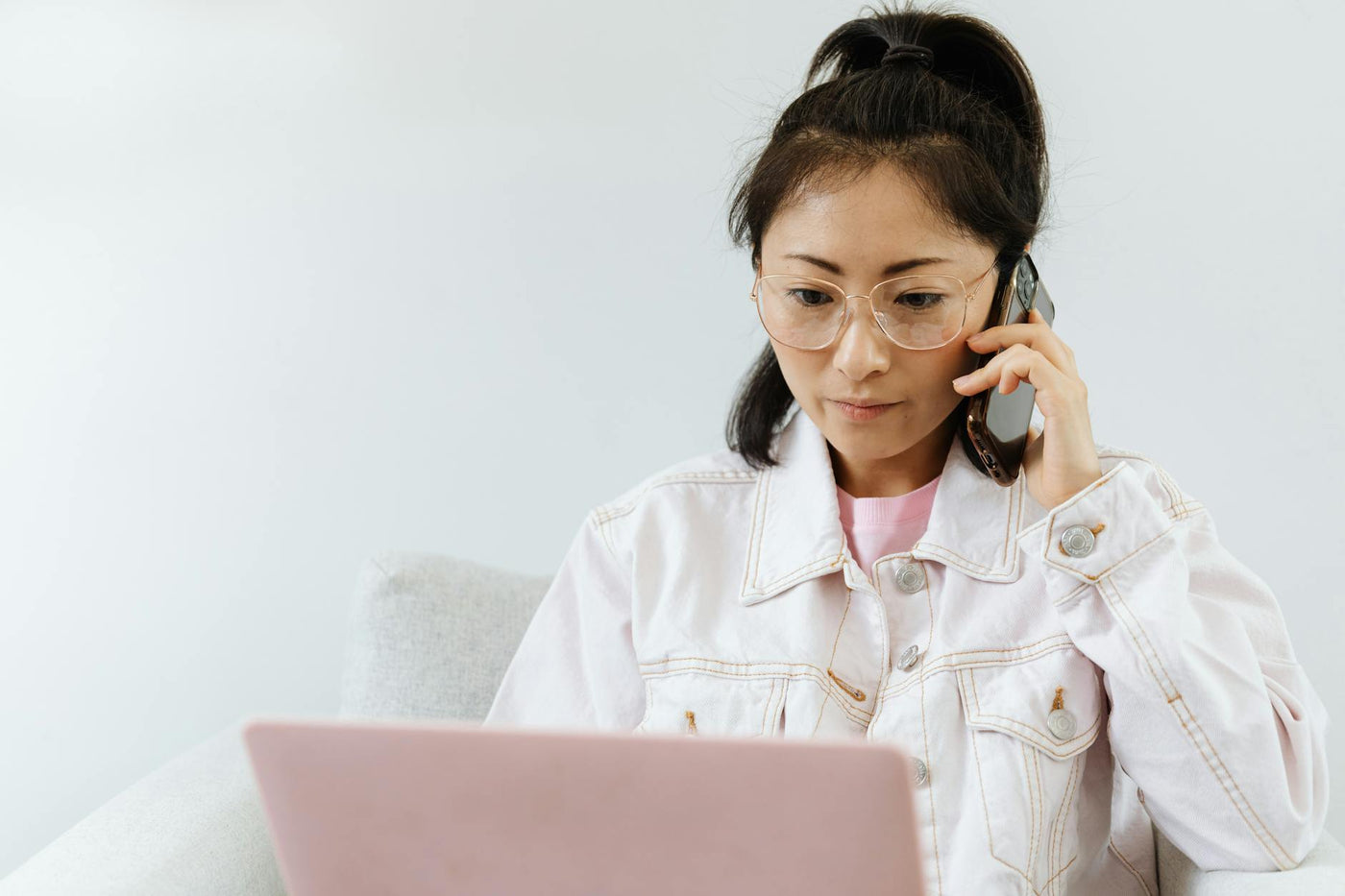 Woman in glasses multitasking with phone and laptop in a serene office setting.