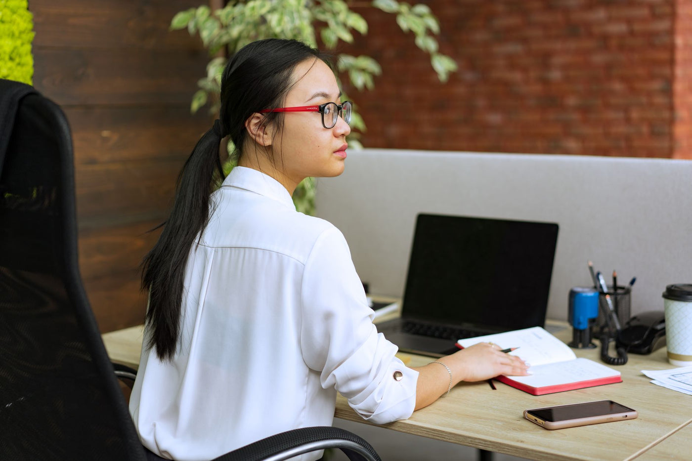 Professional Asian woman in glasses working at a desk with a laptop and notebook in an office.
