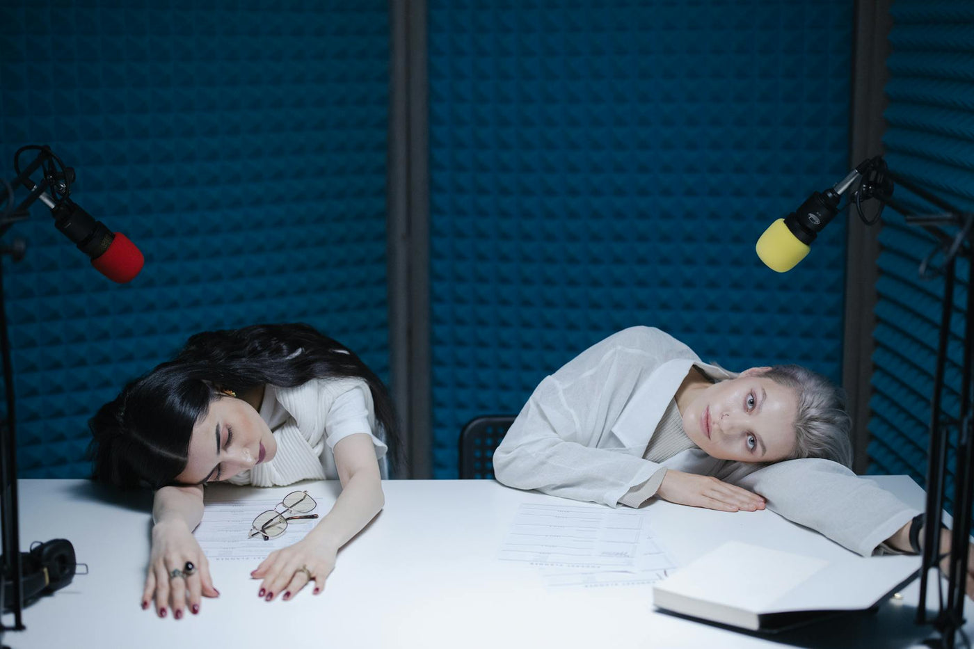 Two women resting on a white table during podcast recording in a soundproof studio.