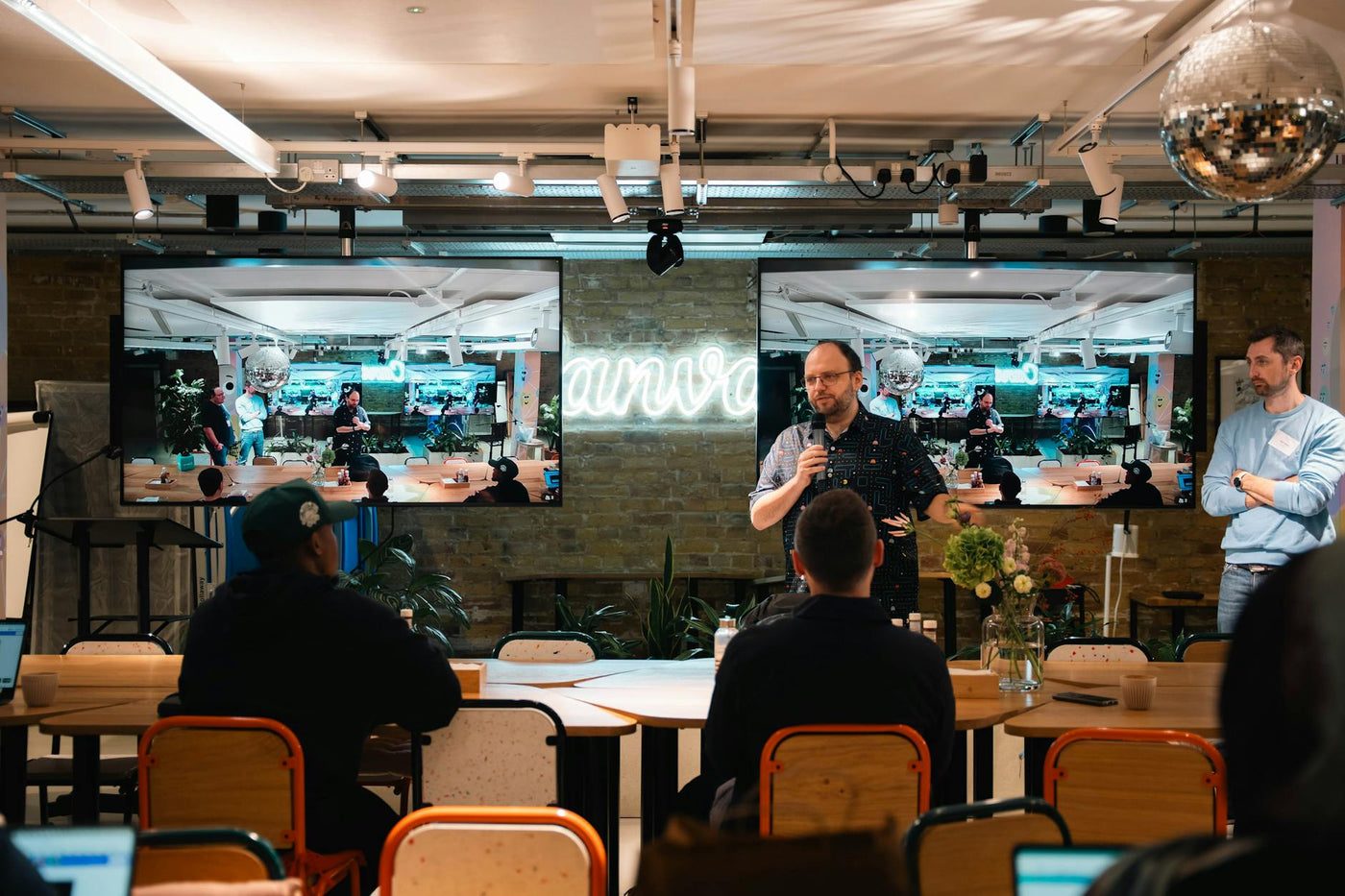 Speaker addresses audience in a modern indoor conference setting, featuring a live presentation on large screens.