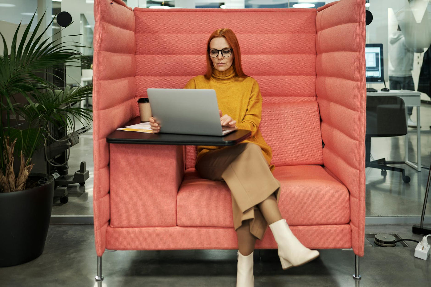Businesswoman in a yellow sweater working on a laptop in a modern office booth setting, exhibiting style and focus.